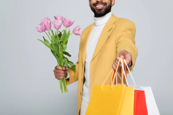 Vista cortada de homem americano africano feliz em blazer amarelo segurando tulipas rosa e sacos de compras isolados em cinza — Fotografia de Stock