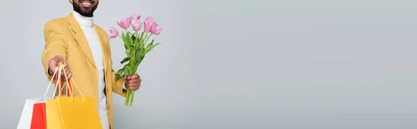 Abgeschnittene Ansicht eines glücklichen afrikanisch-amerikanischen Mannes im gelben Blazer, der rosa Tulpen und Einkaufstaschen isoliert auf grau hält, Banner — Stockfoto