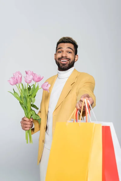 Улыбающийся африканский американец в жёлтом пиджаке и поло с розовыми тюльпанами и пакетами для покупок, изолированными на сером — стоковое фото