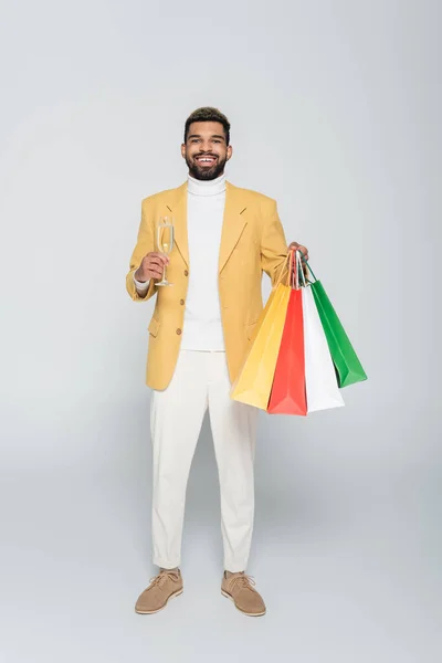 Pleine longueur de heureux homme afro-américain en blazer jaune tenant des sacs à provisions et une coupe de champagne sur gris — Photo de stock