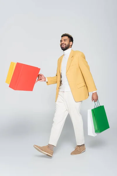 Longitud completa de hombre americano africano alegre en chaqueta amarilla sosteniendo bolsas de compras y caminando sobre gris - foto de stock
