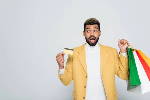 Sorprendido hombre afroamericano en chaqueta amarilla sosteniendo bolsas de compras y tarjeta de crédito aislado en gris - foto de stock