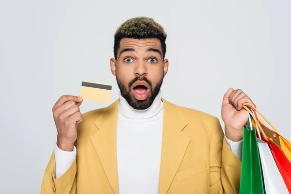 Здивований афроамериканський чоловік у жовтому блістері тримає сумки для покупок та кредитну картку ізольовано на сірому — стокове фото
