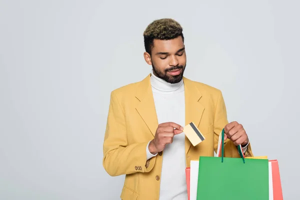 Barbudo hombre afroamericano en chaqueta amarilla sosteniendo bolsas de compras y tarjeta de crédito aislado en gris - foto de stock