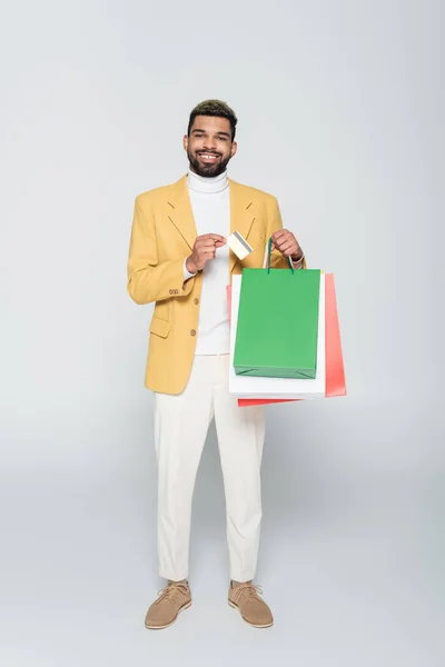 Full length of happy african american man in yellow blazer holding shopping bags and credit card on grey — Stock Photo
