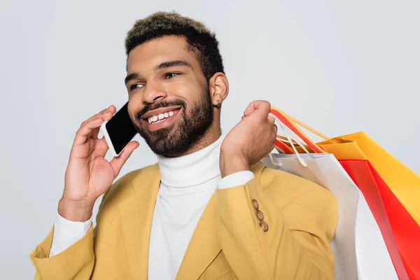 Allegro uomo afroamericano in giacca gialla e collo a polo che tiene borse della spesa e parla sul cellulare isolato su grigio — Foto stock