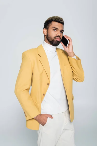 Junger afrikanisch-amerikanischer Mann im gelben Blazer spricht mit Smartphone und posiert mit der Hand in der Tasche isoliert auf grau — Stockfoto