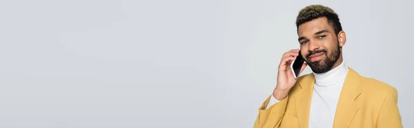 Cheerful african american man in yellow blazer talking on smartphone isolated on grey, banner — Stock Photo