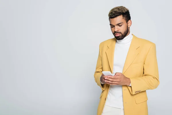 Barbudo hombre afroamericano en blazer amarillo mensajes de texto en el teléfono inteligente aislado en gris - foto de stock