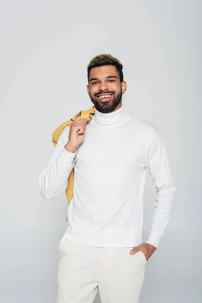 Heureux homme afro-américain avec les yeux bleus à col roulé tenant blazer regardant caméra isolée sur gris — Photo de stock