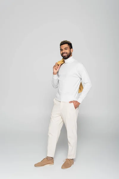 Comprimento total de homem americano africano feliz em gola alta segurando blazer e posando com a mão no bolso em cinza — Fotografia de Stock