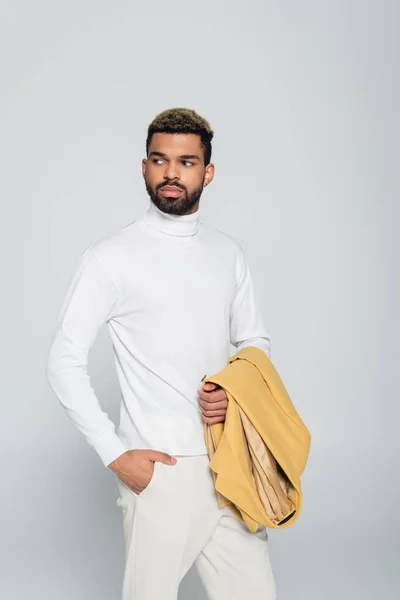 Happy african american man with blue eyes in turtleneck holding blazer and posing with hand in pocket isolated on grey — Stock Photo