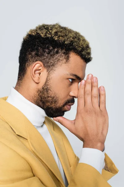 Vista lateral del joven afroamericano en elegante chaqueta amarilla con las manos orantes aisladas en gris - foto de stock