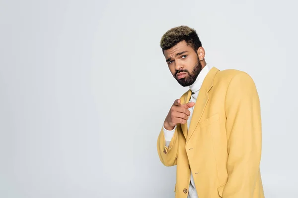 Barbudo hombre afroamericano con ojos azules en elegante chaqueta amarilla apuntando con el dedo aislado en gris - foto de stock