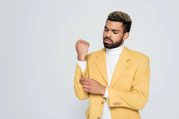 Joven afroamericano hombre con ojos azules en elegante chaqueta amarilla ajustando la manga aislado en gris - foto de stock