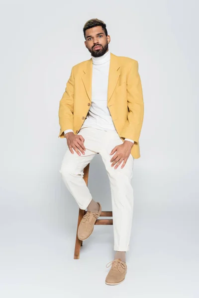 Full length of bearded african american man in stylish outfit sitting on wooden high chair on grey — Stock Photo