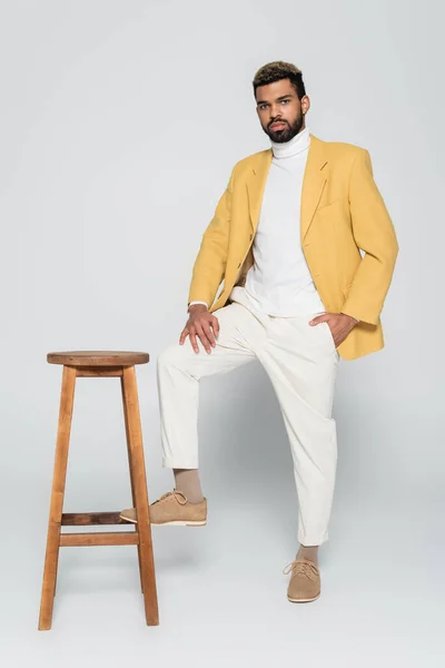 Full length of bearded african american man in stylish outfit standing with hand in pocket near wooden high chair on grey — Stock Photo