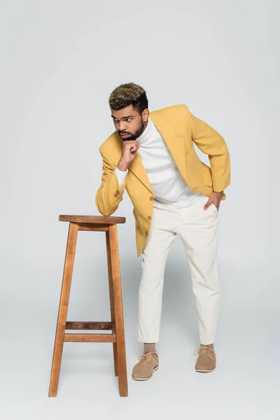 Longitud completa de hombre afroamericano en traje elegante de pie con la mano en el bolsillo cerca de la silla alta de madera en gris - foto de stock