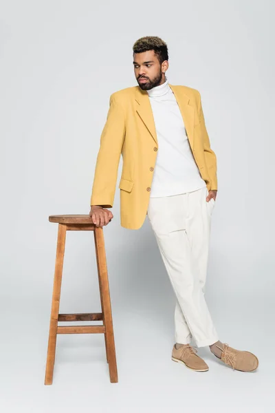 Повна довжина бородатого афроамериканця в стильному вбранні, що стоїть з рукою в кишені біля дерев'яного високого стільця на сірому — стокове фото