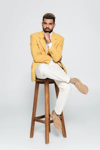 Full length of bearded african american man in stylish outfit sitting on wooden high chair on grey — Stock Photo