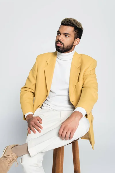 Bearded african american man in stylish outfit sitting on wooden high chair isolated on grey — Stock Photo