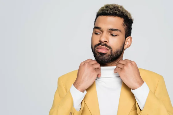 Barbudo hombre afroamericano en elegante chaqueta ajustando cuello de polo aislado en gris - foto de stock