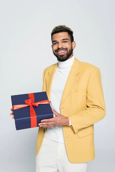 Heureux homme afro-américain en jaune veston élégant tenant présent enveloppé isolé sur gris — Photo de stock