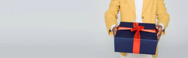 Vista recortada del hombre afroamericano en blazer elegante amarillo sosteniendo regalo envuelto aislado en gris, bandera - foto de stock