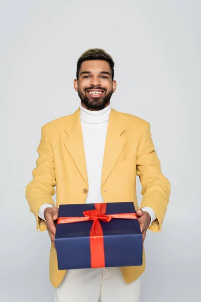 Alegre africano americano hombre en amarillo elegante chaqueta celebración envuelto presente aislado en gris - foto de stock