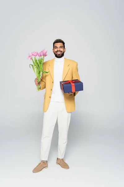 Full length of happy african american man in yellow blazer holding wrapped present and tulips on grey — Stock Photo