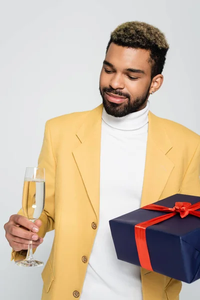 Bärtiger afrikanisch-amerikanischer Mann in gelbem, stylischem Blazer mit umwickeltem Geschenk und Glas Champagner isoliert auf grau — Stockfoto