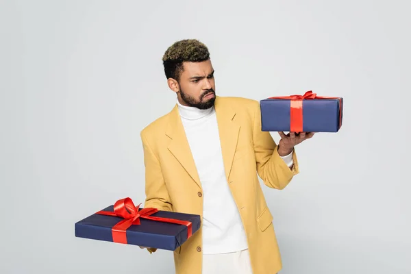 Curioso africano americano hombre en amarillo elegante chaqueta celebración envuelto regalos aislado en gris - foto de stock