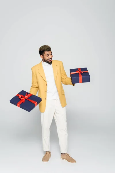 Full length of excited african american man in yellow stylish blazer holding wrapped presents on grey — Stock Photo