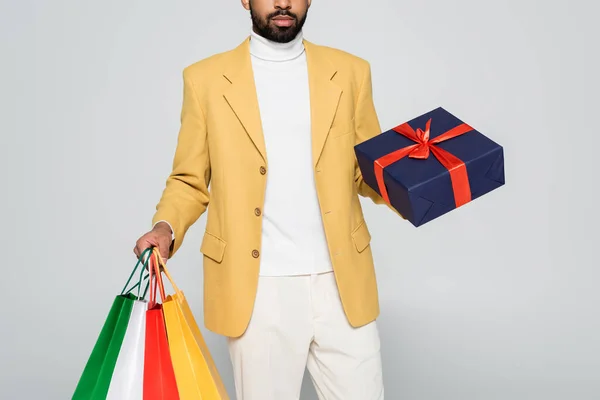Vista cortada do homem americano africano barbudo em blazer amarelo segurando presente e sacos de compras isolados em cinza — Fotografia de Stock