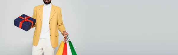 Vista recortada del hombre afroamericano barbudo en chaqueta amarilla sosteniendo presente y bolsas de compras aisladas en gris, pancarta - foto de stock