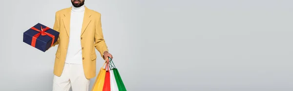 Cropped view of bearded african american man in yellow blazer holding present and shopping bags isolated on grey, banner — Stock Photo