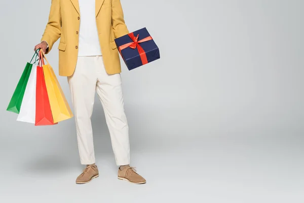 Vista recortada del hombre afroamericano en blazer elegante amarillo sosteniendo regalo envuelto y bolsas de compras aisladas en gris - foto de stock