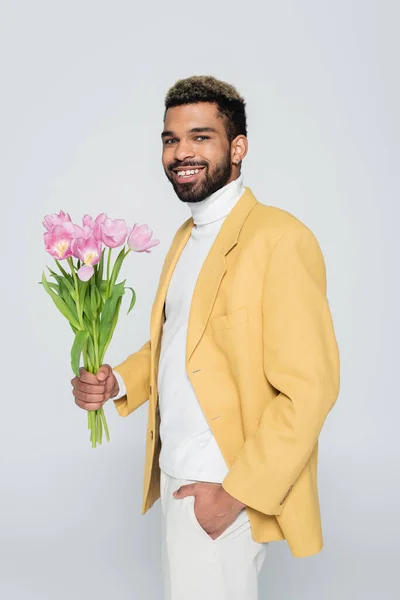 Happy african american man in stylish blazer holding bouquet of pink tulips isolated on grey — Stock Photo