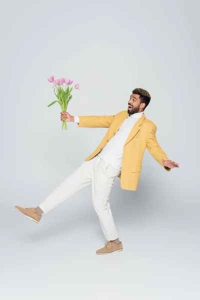 Full length of exited african american man in stylish blazer holding bouquet of pink tulips on grey — Stock Photo