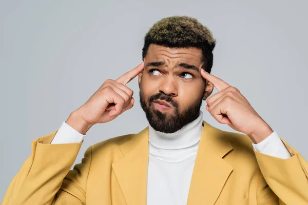 Joven afroamericano hombre en elegante chaqueta apuntando a los templos y pensando aislado en gris - foto de stock