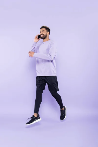 Full length of happy african american man talking on smartphone and levitating on purple — Stock Photo