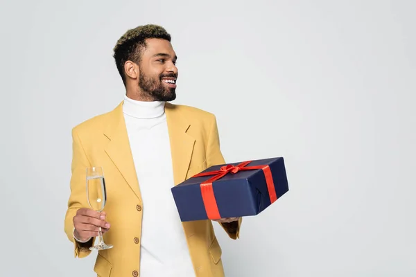 Homme afro-américain souriant en blazer jaune tenant présent enveloppé et verre de champagne isolé sur gris — Photo de stock