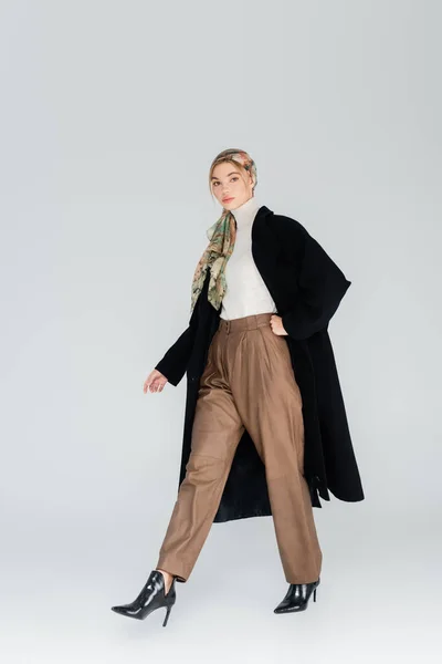 Pleine longueur de femme élégante en manteau noir et foulard marchant sur fond gris — Photo de stock