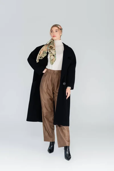 Longueur totale de la femme en manteau noir et pantalon beige posant avec la main à la taille sur fond gris — Photo de stock
