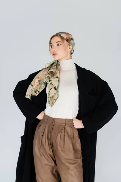 Jeune femme en foulard et manteau noir debout avec les mains à la taille et regardant loin isolé sur gris — Photo de stock