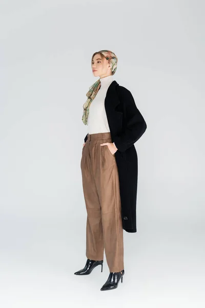 Full length view of stylish woman in black coat standing with hands in pockets of beige trousers on grey background — Stock Photo