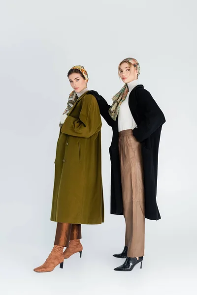 Full length of women in elegant kerchiefs and coats looking at camera on grey background — Stock Photo