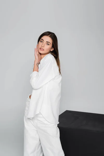 Brunette woman in white shirt touching face near black cube isolated on grey — Stock Photo