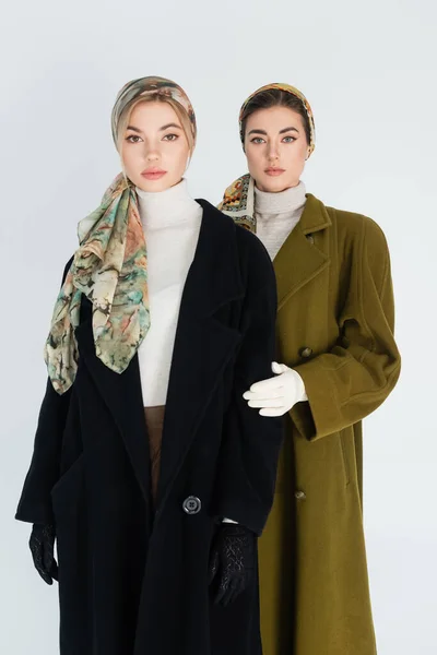 Mujeres elegantes en pañuelos para la cabeza y abrigos mirando a la cámara aislada en gris - foto de stock