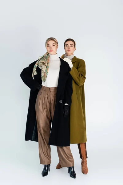 Piena lunghezza di donne eleganti in fazzoletti e cappotti guardando la fotocamera su sfondo grigio — Foto stock